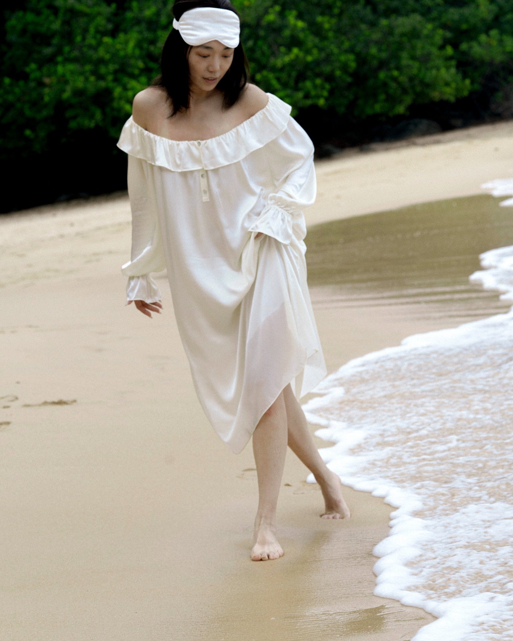 Woman walking barefoot on the beach wearing an off-shoulder white mulberry silk nightdress and matching silk sleep mask by Liu&Liu – luxury silk sleepwear for coastal retreats.