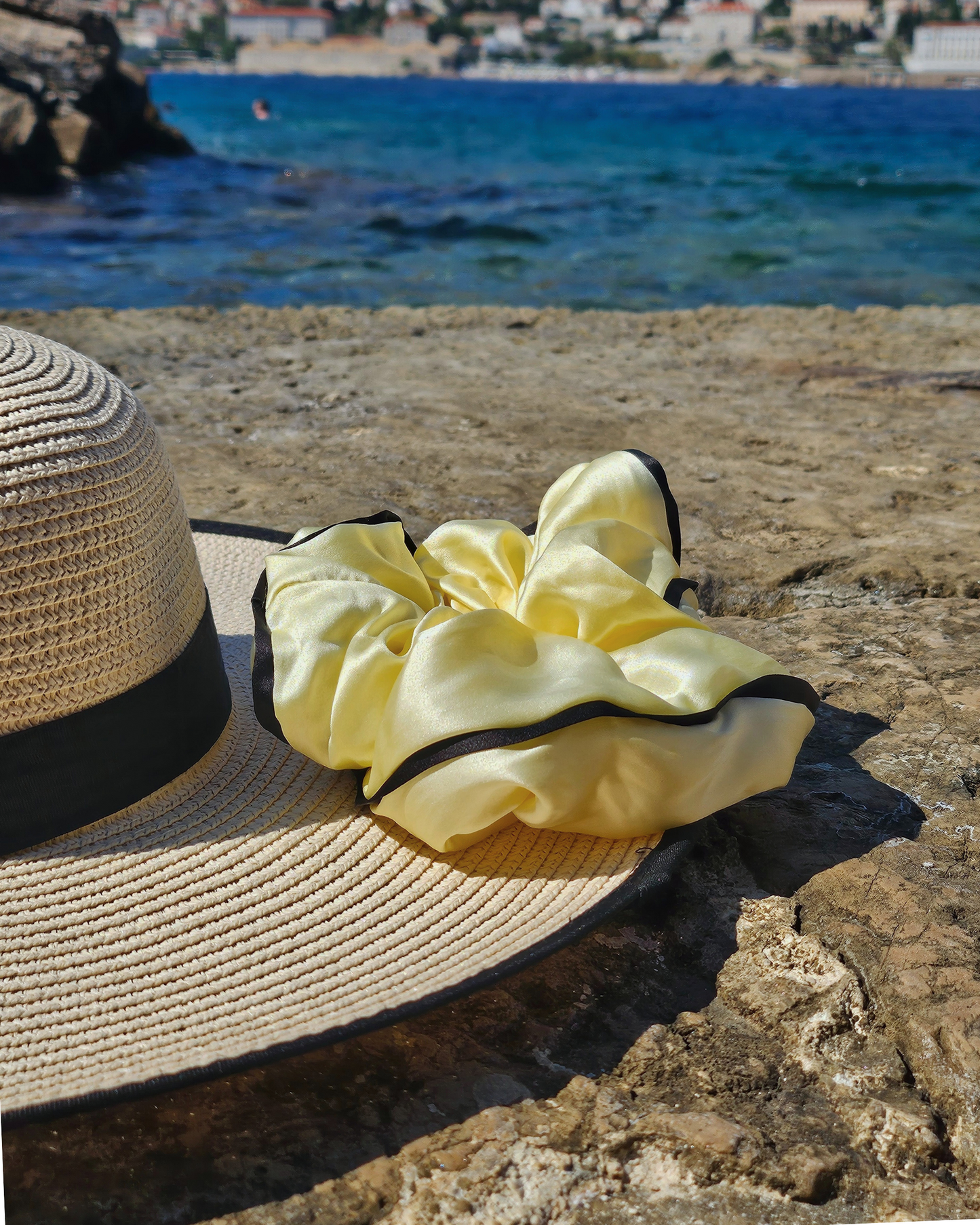Yellow mulberry silk scrunchie with black trim from Liu&Liu resting on a straw sun hat on a rocky beach, with blue sea and a coastal town in the background – luxury silk hair accessory for summer holidays.