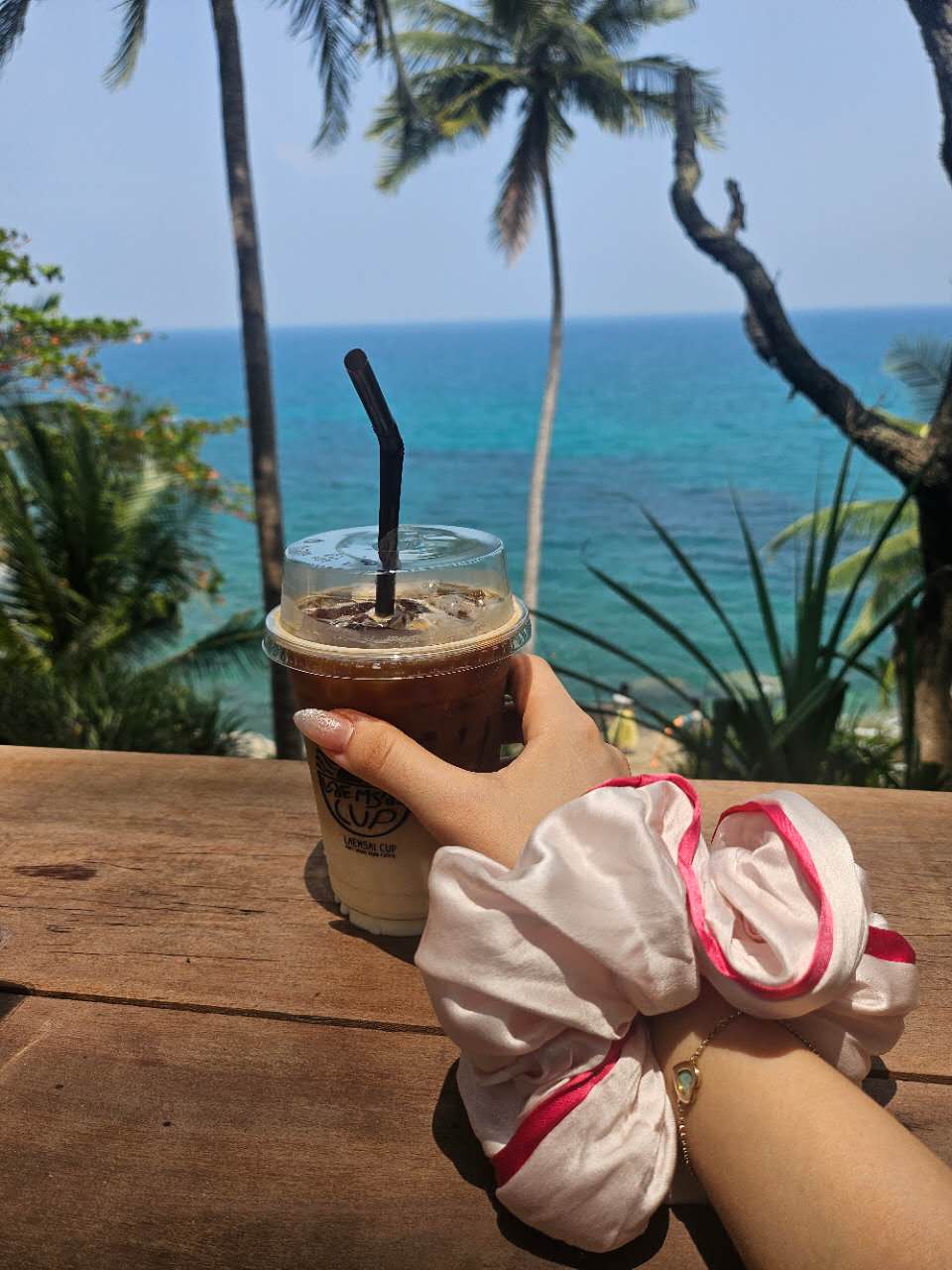 Hand holding an iced coffee by the ocean, wearing a white mulberry silk scrunchie with pink trim from Liu&Liu – luxury silk hair accessory styled for tropical beach holidays.