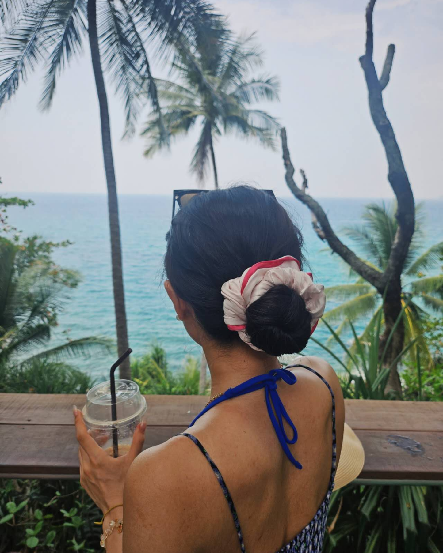 Woman with hair in a bun styled with a white and pink mulberry silk scrunchie by Liu&Liu, holding an iced drink and looking out at a tropical ocean view with palm trees – luxury silk hair accessory for beach holidays.