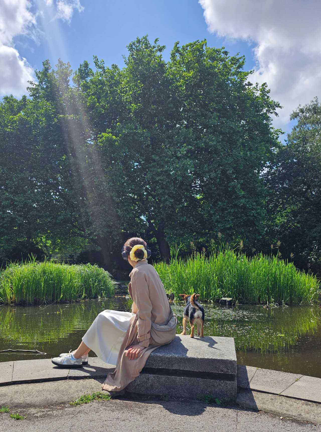 Woman wearing a yellow mulberry silk scrunchie by Liu&Liu, styled in her hair while sitting by a pond with a small dog, sunlit park background – luxury silk hair accessory for everyday elegance.