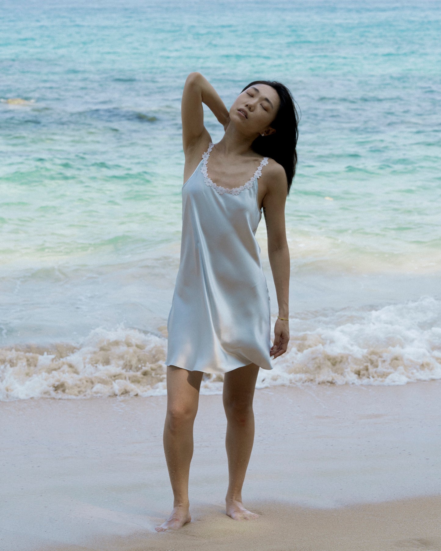 Woman wearing a light blue mulberry silk slip dress with delicate floral spaghetti straps, photographed from the back against the ocean – Liu&Liu luxury silk mini dress for elegant summer style.